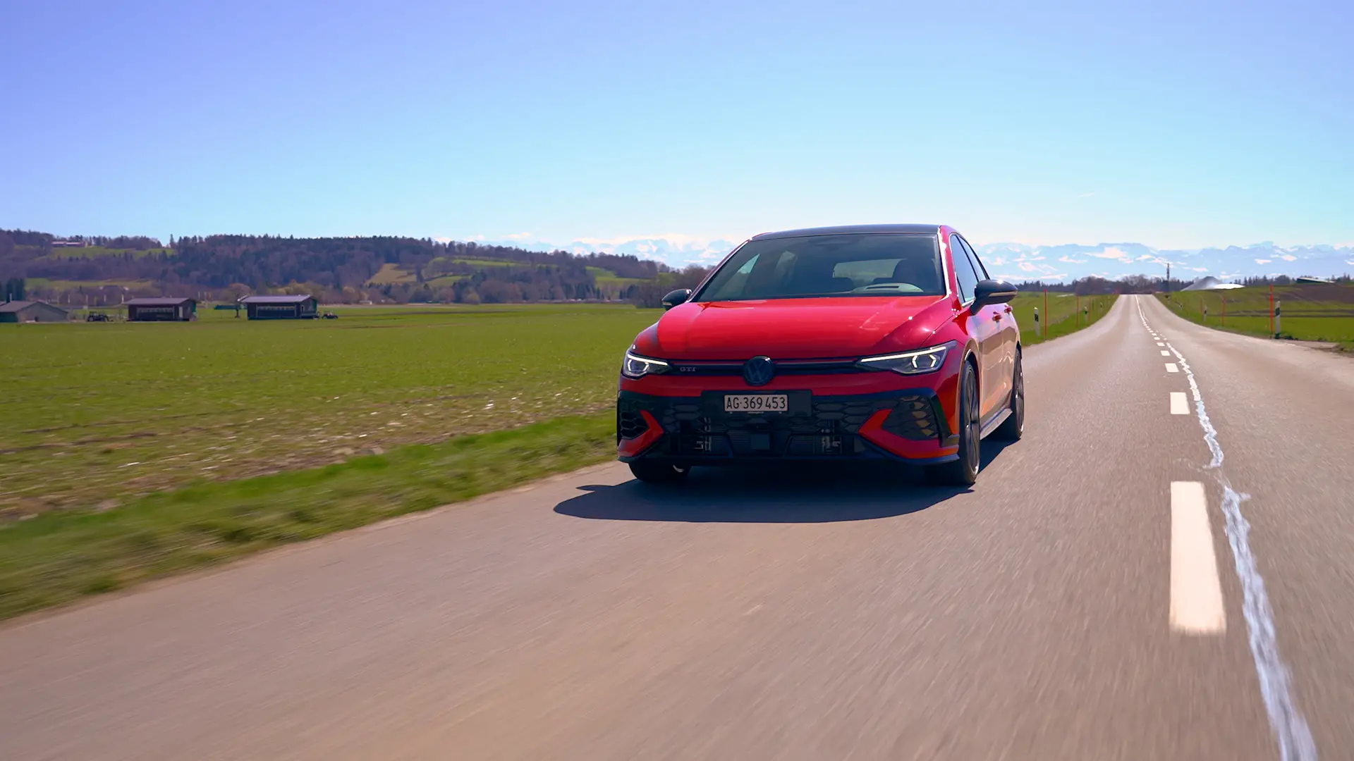 Roter VW Golf fährt auf einer Landstrasse durch offene Felder, im Hintergrund Berge.