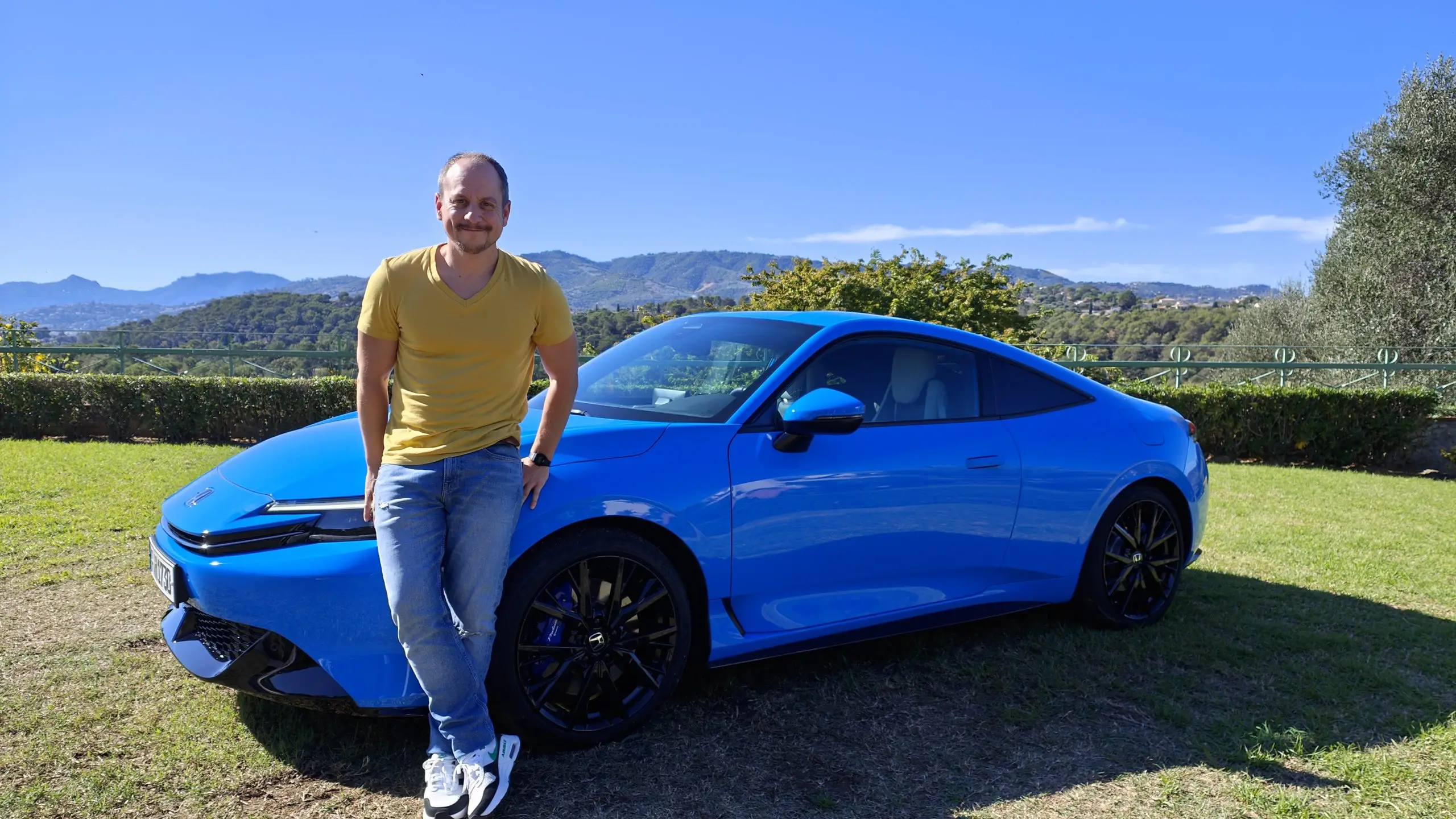 Sämi Pfister lehnt an einem blauen Honda Prelude auf einer Wiese vor Bergkulisse.