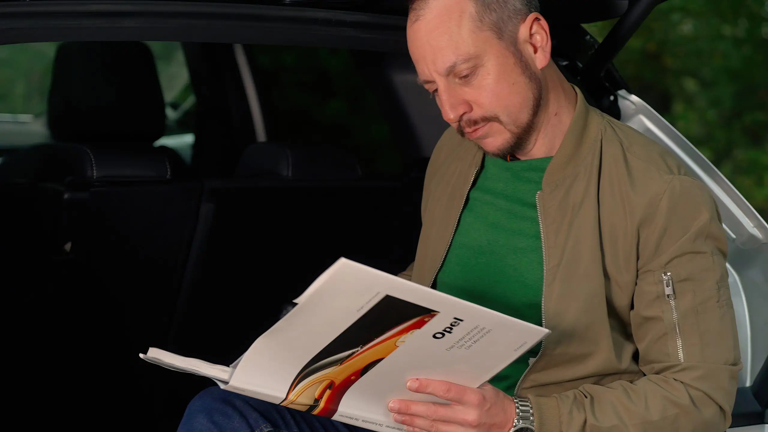 Mann liest in einem aufgeschlagenen Buch über Opel am offenen Kofferraum eines Autos.