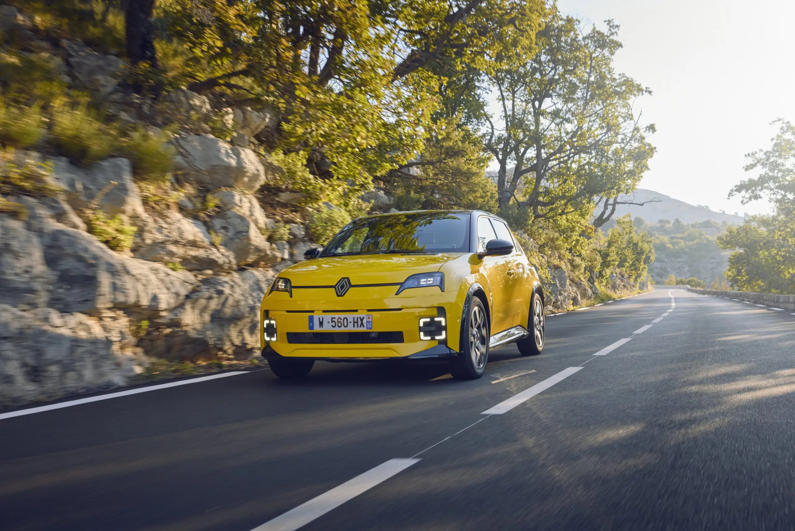 Gelber Renault 5 E-Tech Electric fährt auf kurviger Landstrasse durch felsige Landschaft.