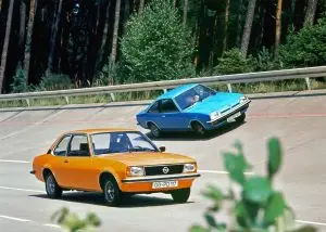 Zwei klassische Opel-Coupés, orange und blau, fahren auf einer kurvigen Strasse am Waldrand.