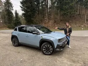 Hellblauer Renault 4 E-Tech Electric auf Waldparkplatz, daneben eine Frau, die auf das Auto zeigt.