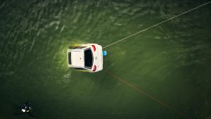 Weisses Testauto treibt im grünen Wasser, gesichert mit Leinen; ein Taucher nähert sich.