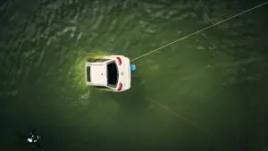 Weisses Testauto treibt im grünen Wasser, gesichert mit Leinen; ein Taucher nähert sich.