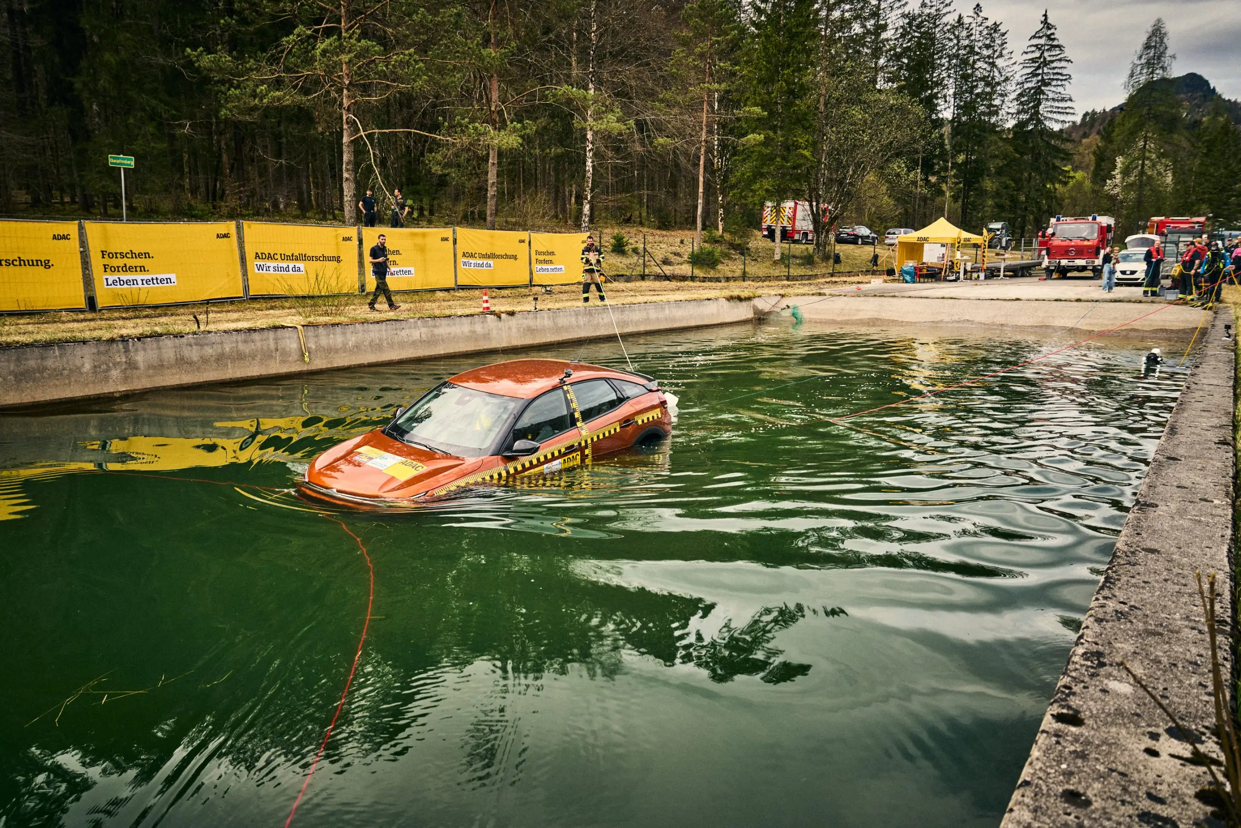 Orangefarbenes Testauto treibt in einem Wasserbecken, Rettungskräfte und Einsatzfahrzeuge stehen am Ufer.