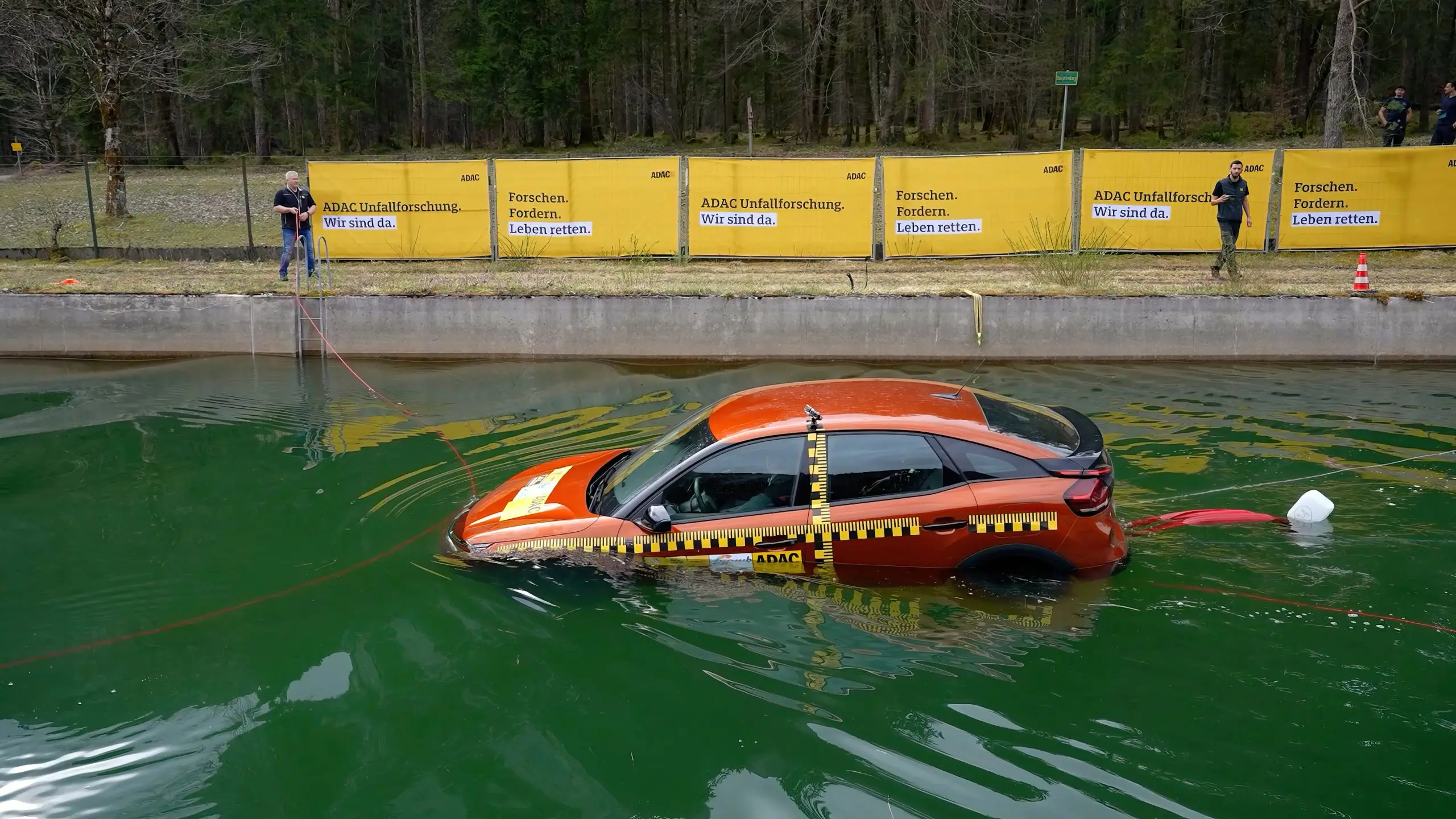 Rotes Testauto steht halb im Wasserbecken, am Ufer beobachten Helfer den Versuch.