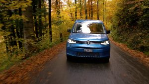 Blauer VW Caddy fährt frontal auf schmaler Waldstrasse im Herbst.
