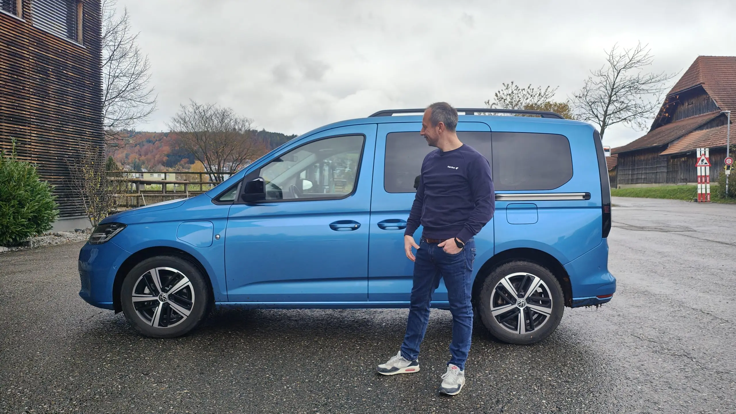 Blauer VW Caddy in Seitenansicht mit Mann daneben auf nassem Hof