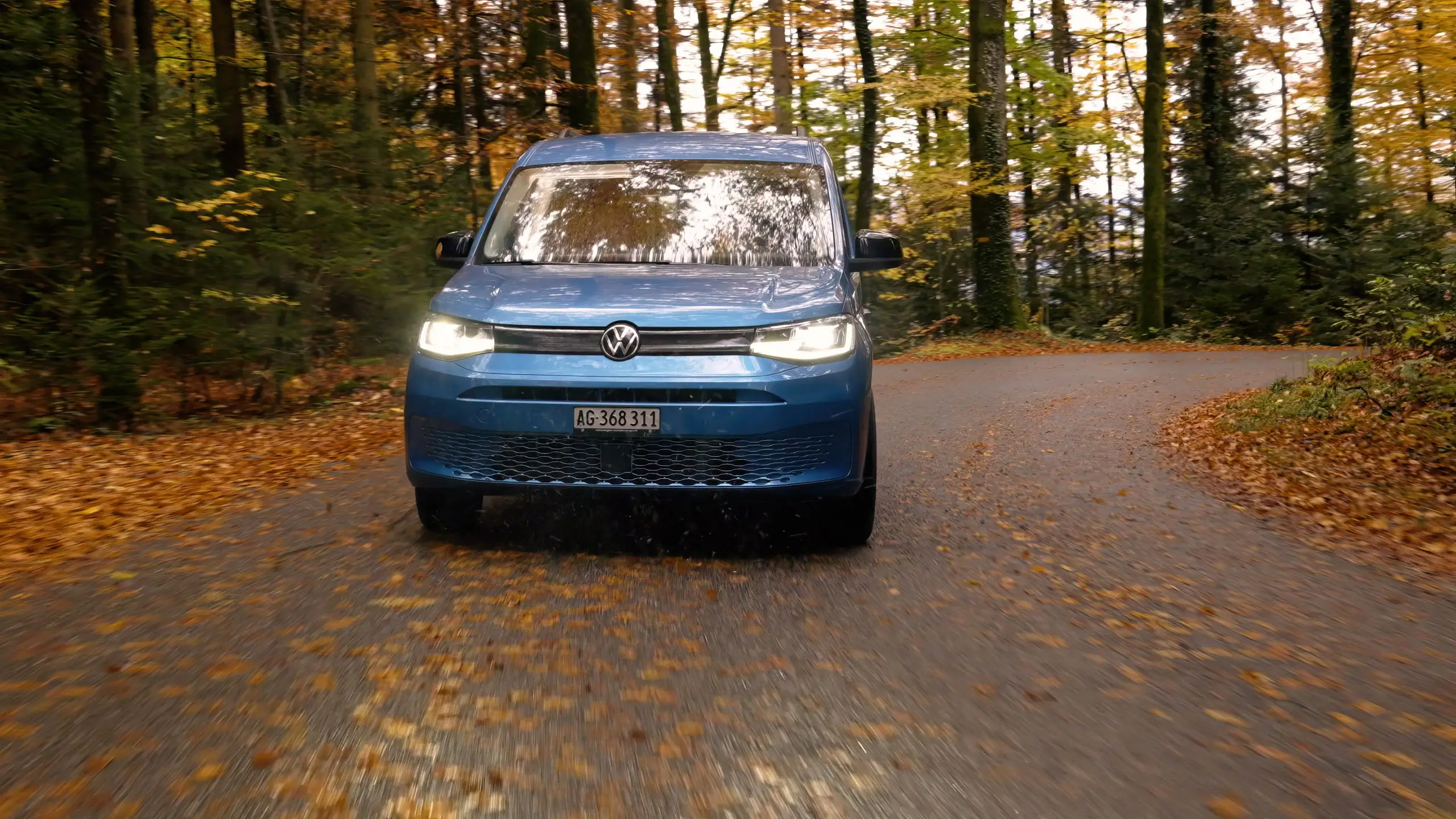 Blauer VW Caddy fährt mit Licht auf herbstlicher Waldstrasse.