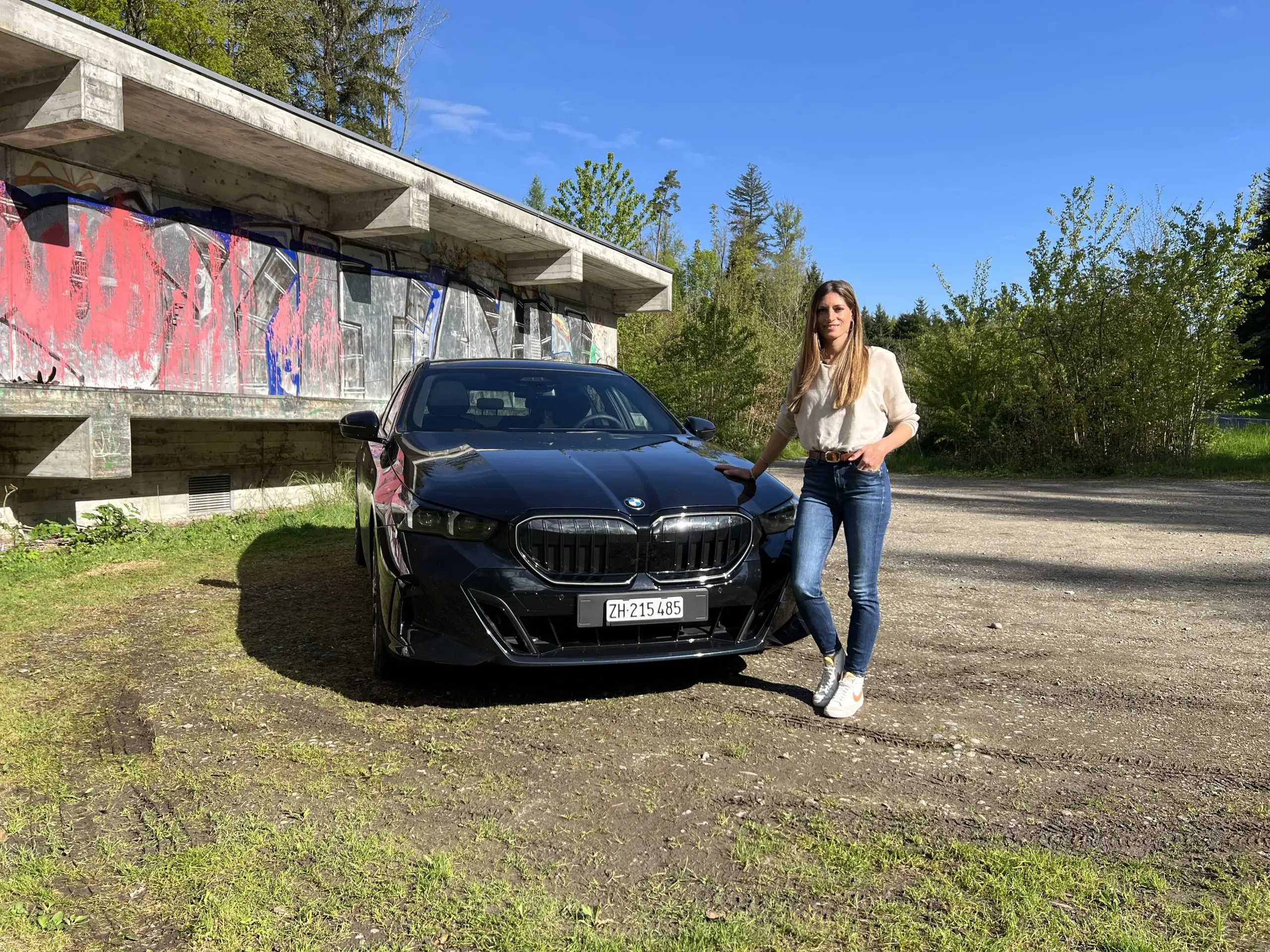 Cyndie Allemann steht neben dem schwarzen BMW 540d auf einem Kiesplatz vor einem Graffiti-Gebäude.