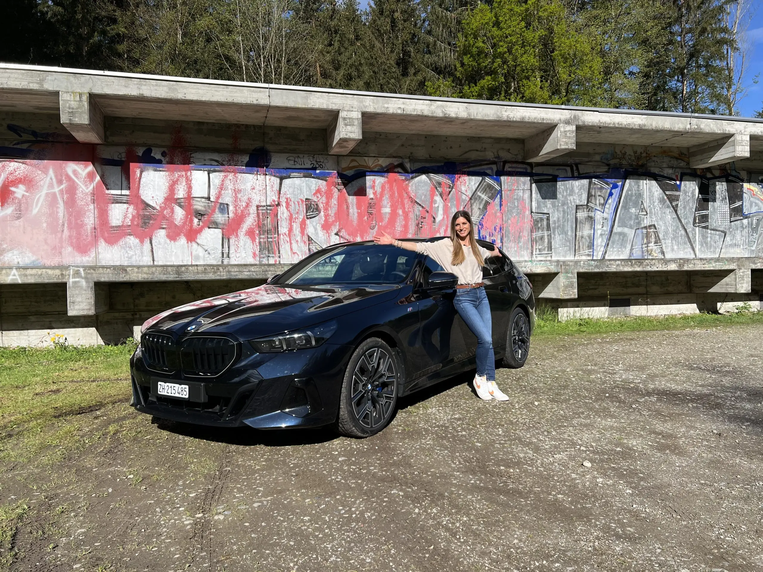 Cyndie Allemann lehnt an einem schwarzen BMW 540d vor einer mit Graffiti besprühten Betonfassade.