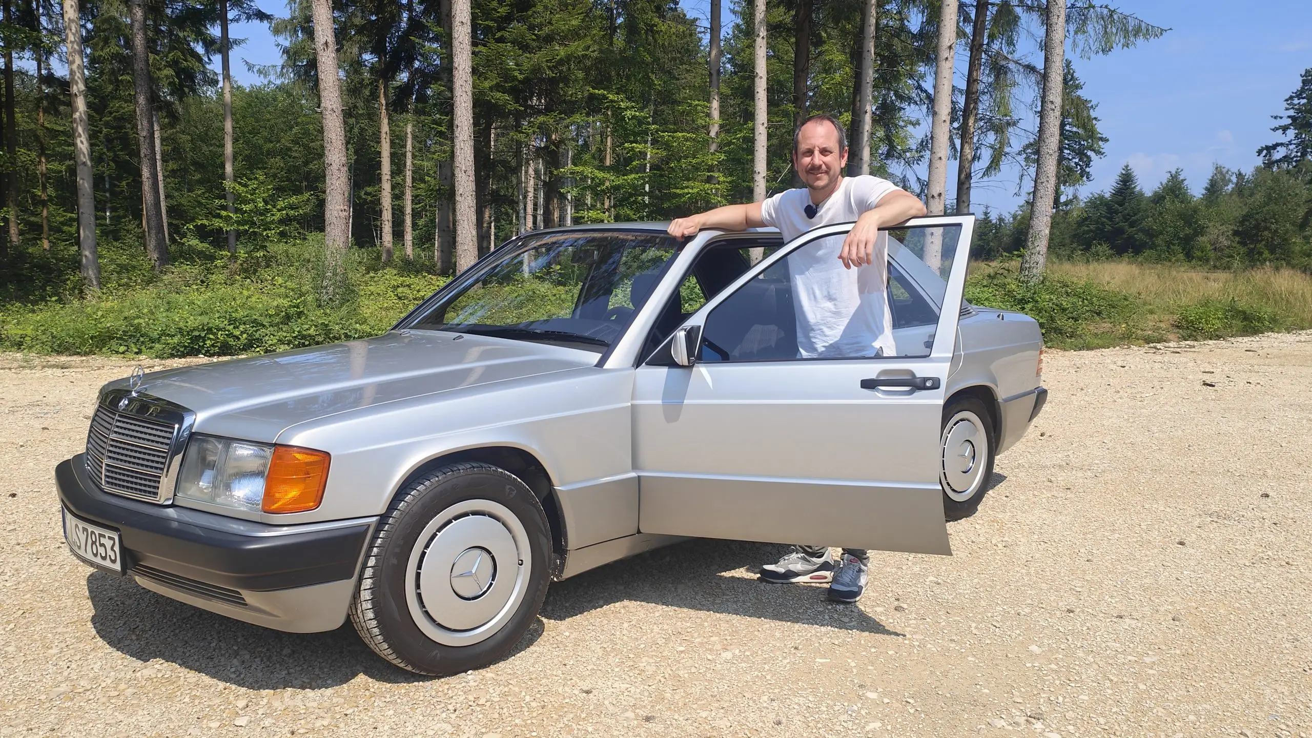 Ein Mann steht neben der geöffneten Fahrertür eines silbernen Mercedes 190 E auf einem Waldweg.