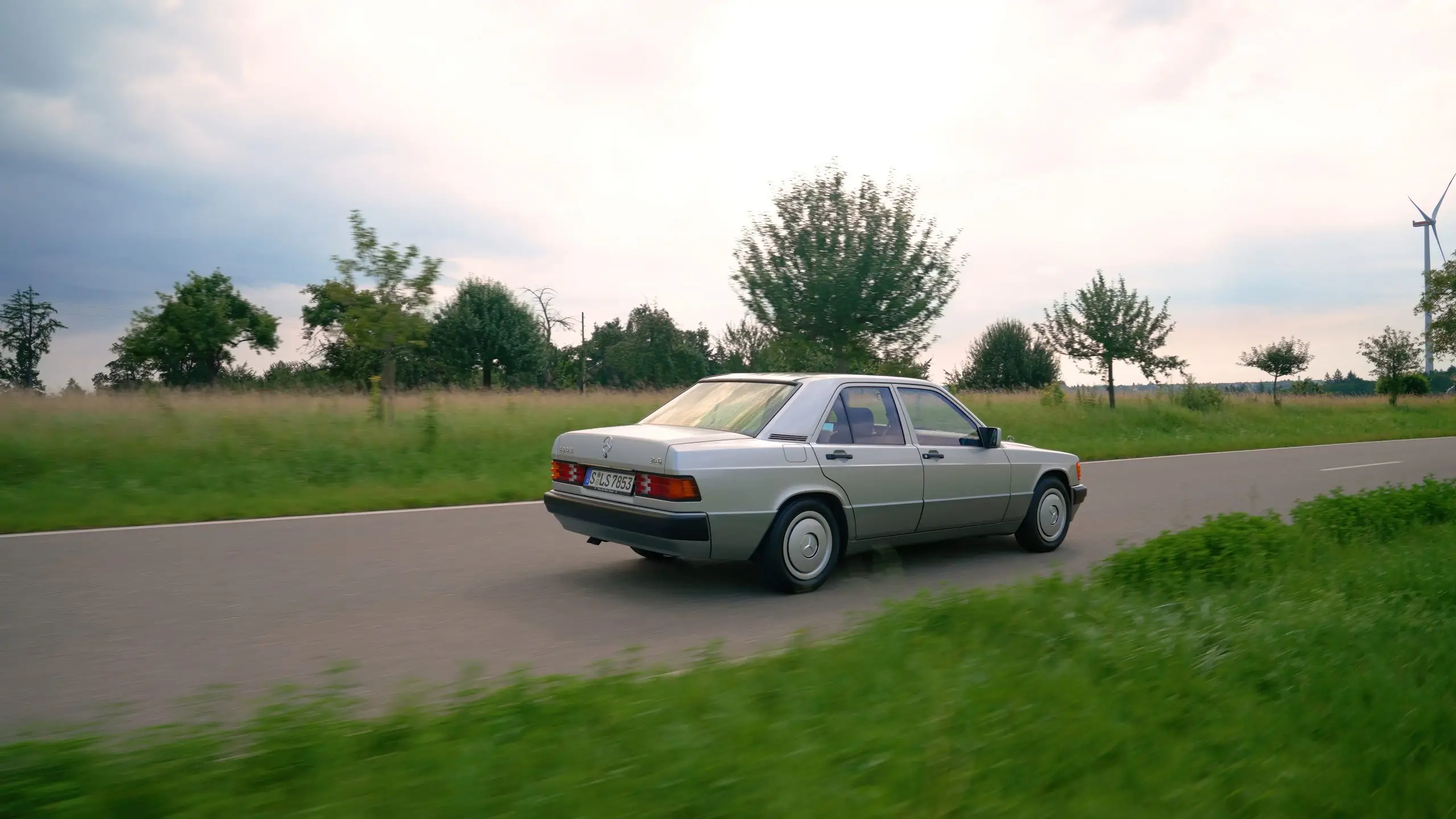 Ein silberner Mercedes 190 E fährt auf einer schmalen Strasse durch eine grüne Landschaft.