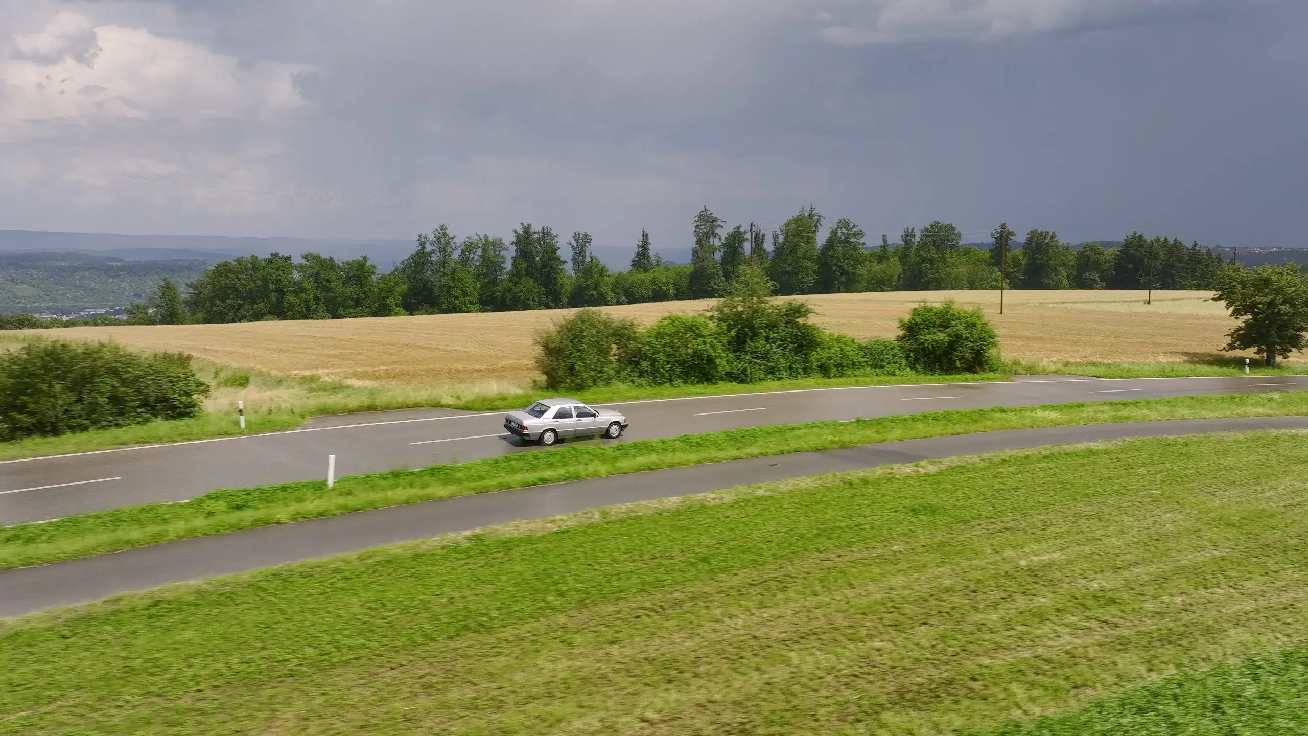 Ein silberner Mercedes 190 E fährt unter dunklen Wolken durch eine offene Feldlandschaft.