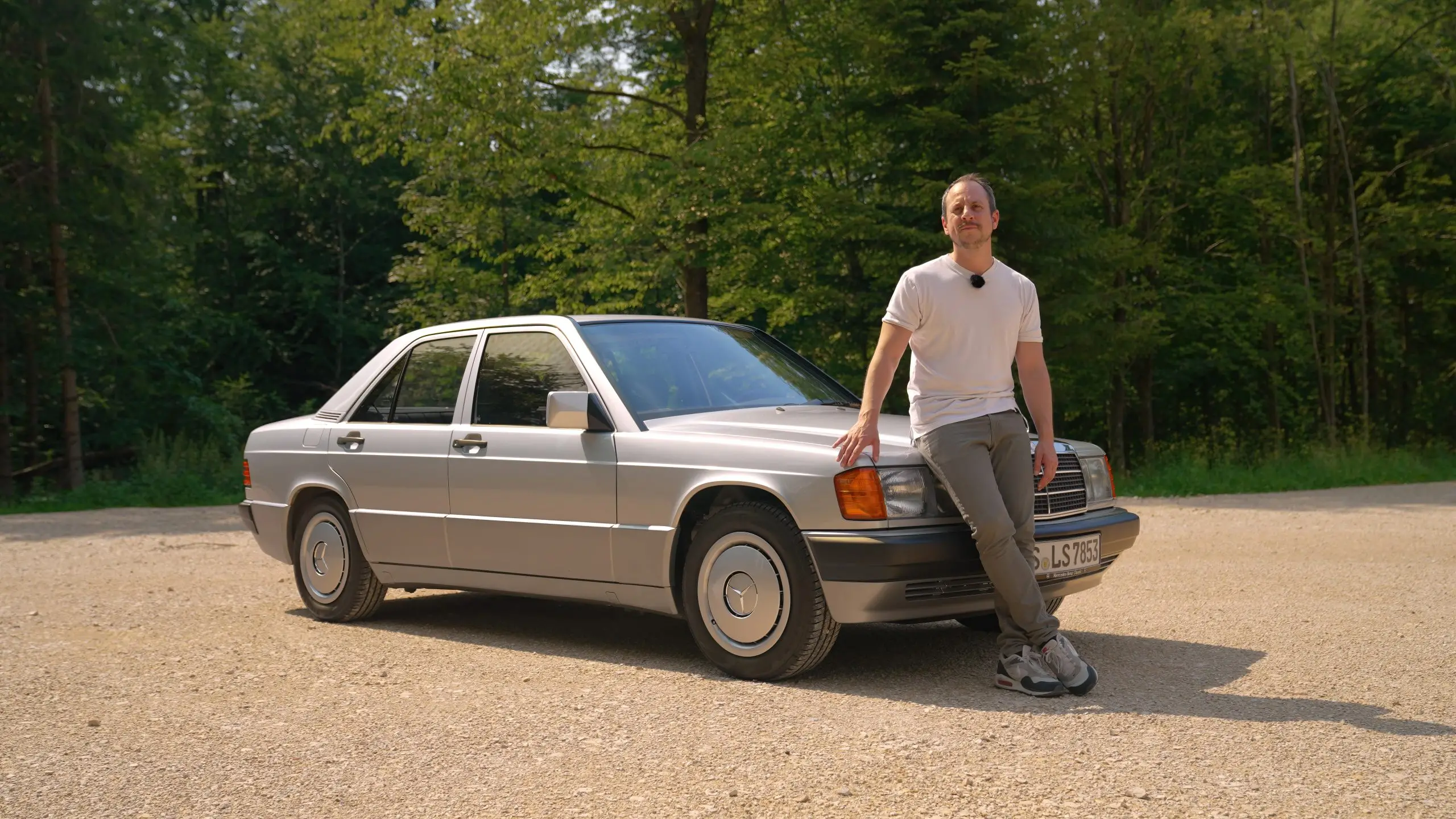 Ein Mann lehnt auf einem Waldparkplatz an der Front eines silbernen Mercedes 190 E.