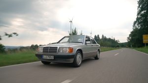 Ein silberner Mercedes 190 E fährt auf einer Landstrasse an Windrädern vorbei.
