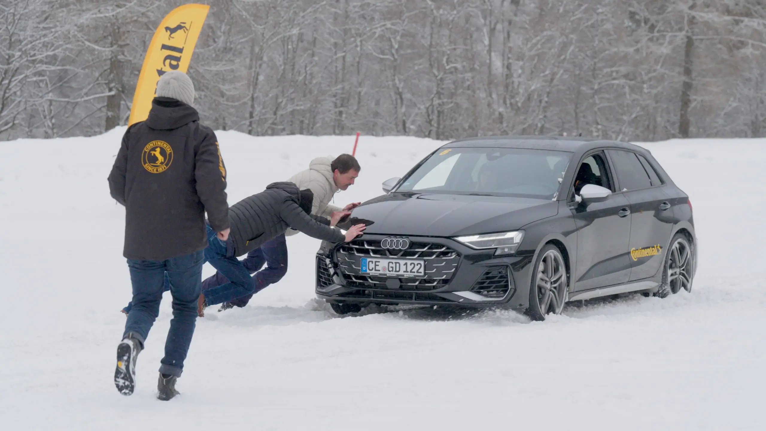 Drei Männer schieben ein schwarzes Auto mit Sommerreifen durch tiefen Schnee.