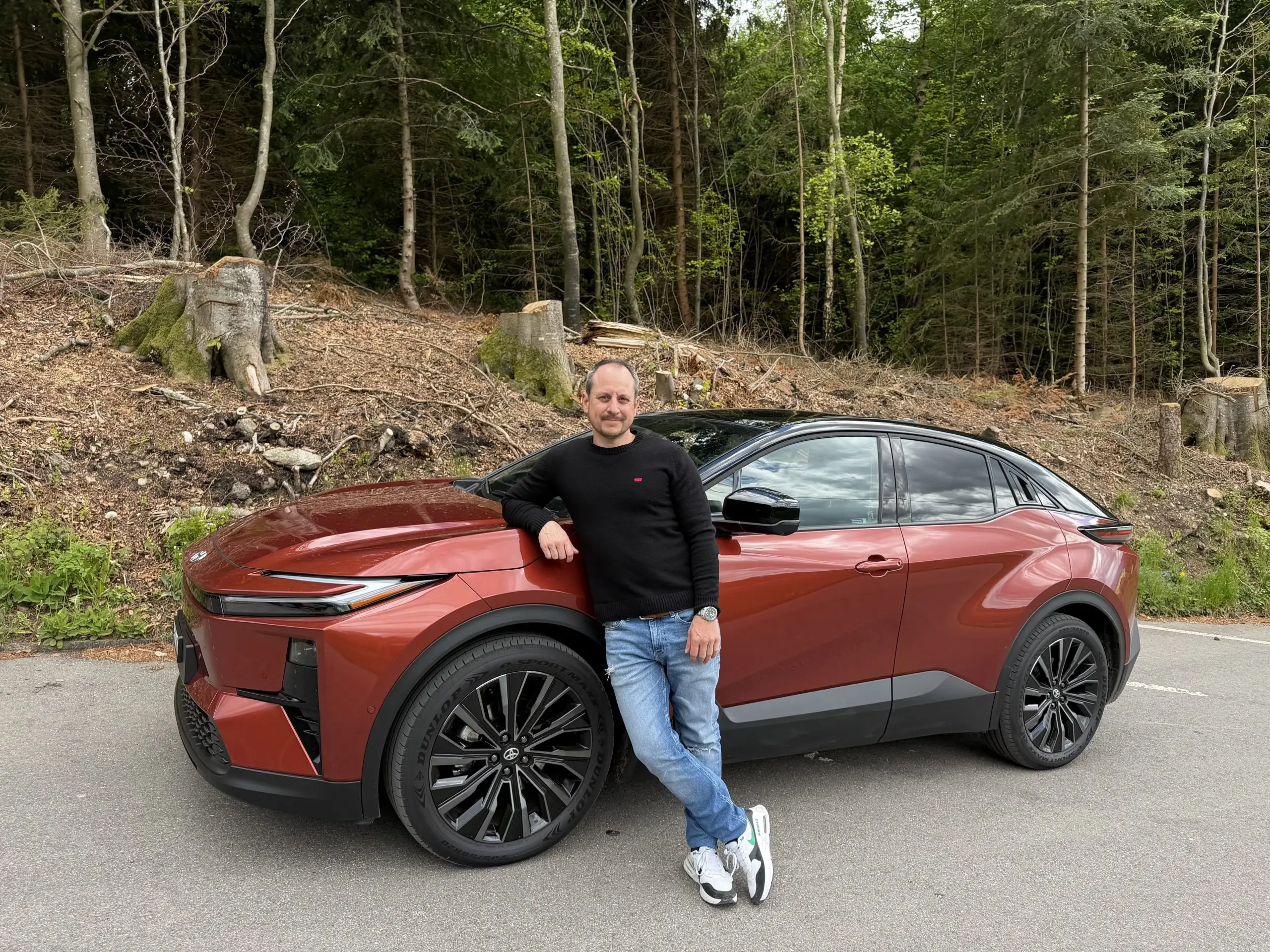 Sämi lehnt an einem roten Toyota C-HR mit schwarzem Dach auf einer Strasse am Waldrand