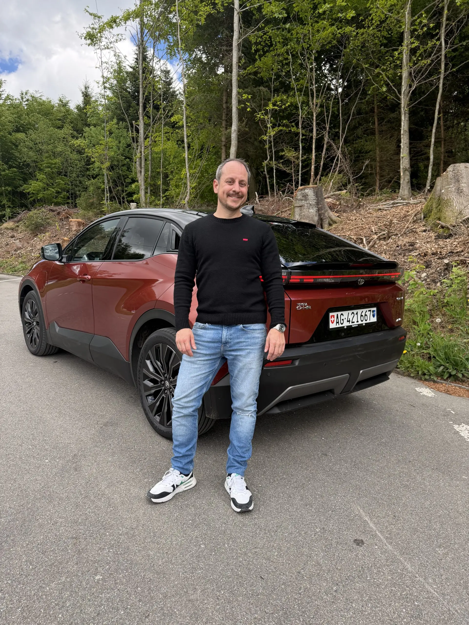 Sämi steht vor einem roten Toyota C-HR+ auf einer Strasse am Waldrand